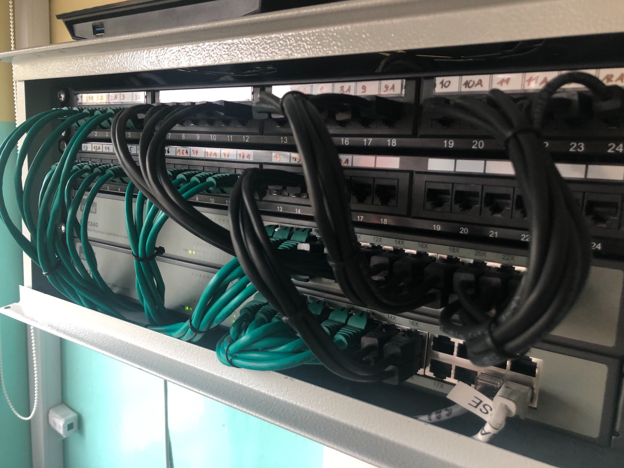 Panel krosowniczy z zielonymi i czarnymi kablami sieciowymi, oznaczony numerami portów, w metalowej szafce na tle turkusowej ściany.