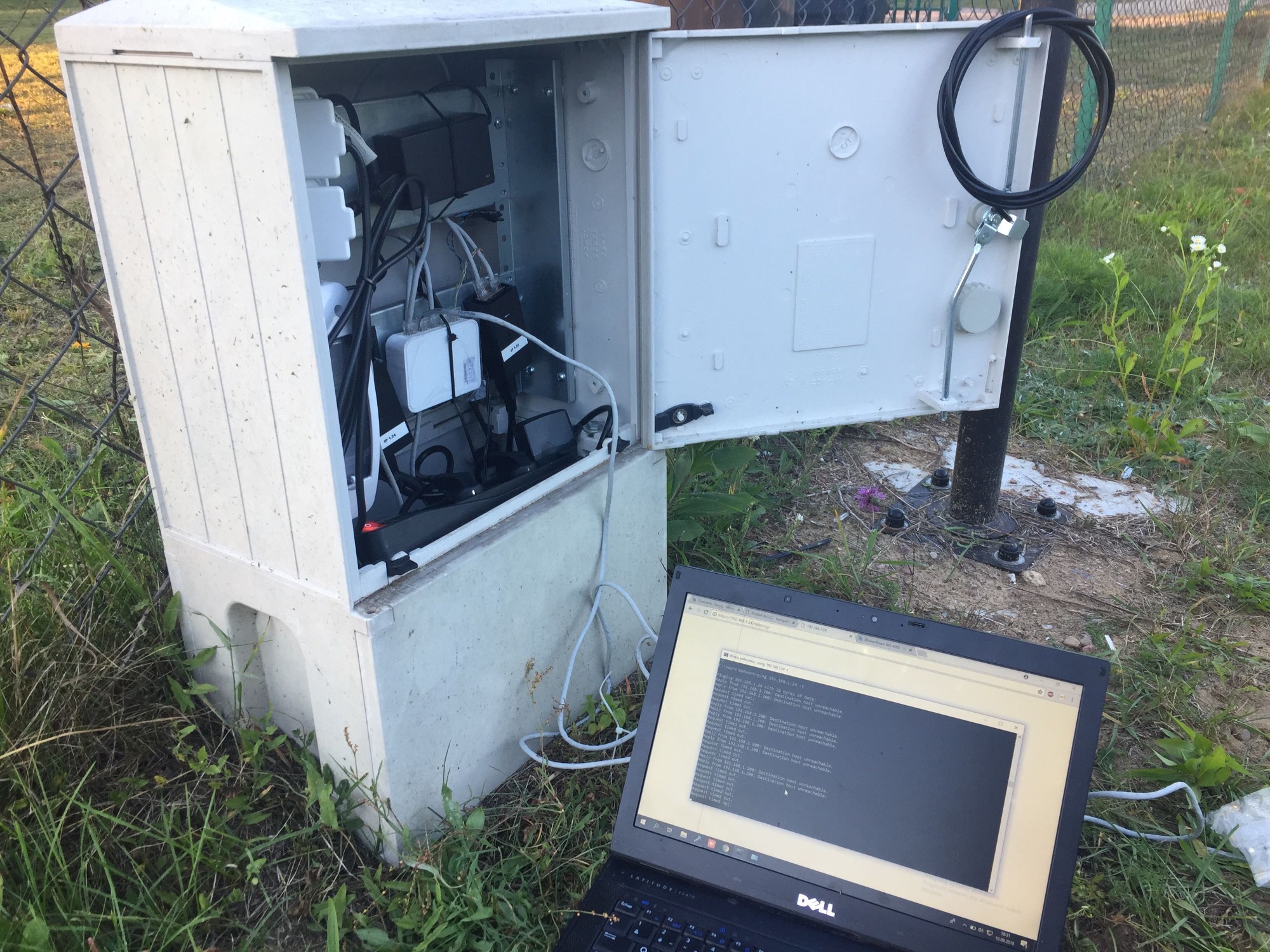 Otwarta szafka telekomunikacyjna z widocznymi kablami i urządzeniami, obok niej laptop Dell z uruchomionym terminalem diagnostycznym, otoczenie to trawa i ogrodzenie.