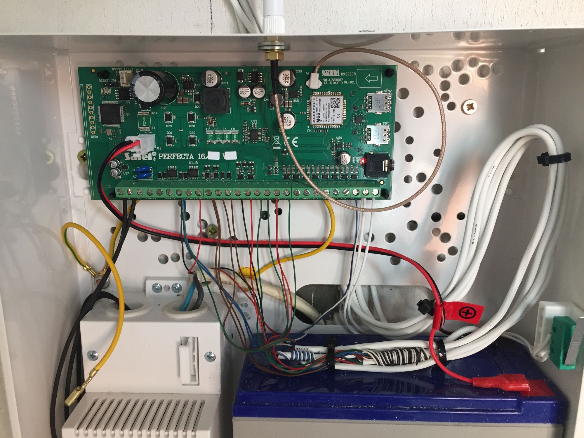 Wnętrze skrzynki alarmowej z płytą główną Satel Perfecta 16, akumulatorem i okablowaniem, widoczne złącza SIM, antena i elementy elektroniczne.
