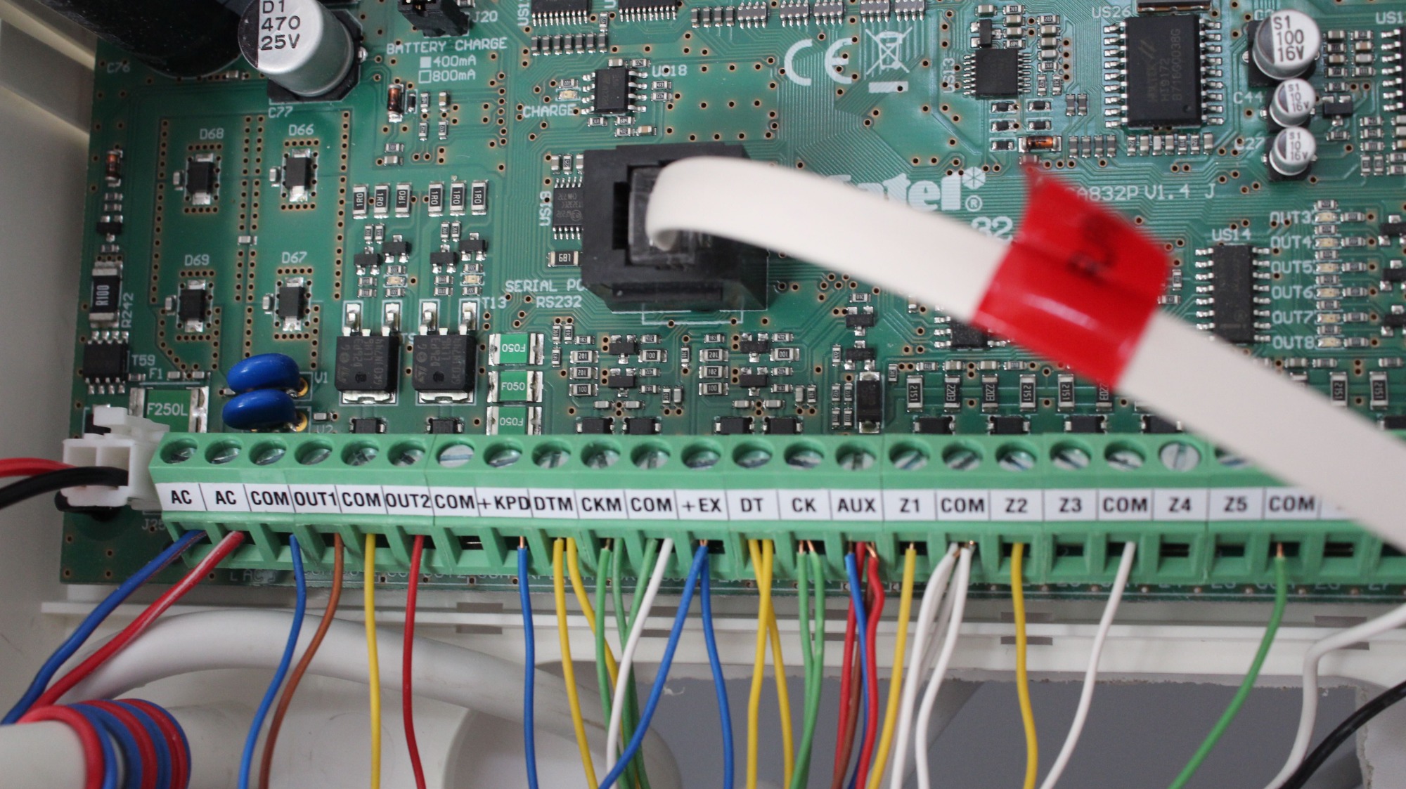 Płyta główna systemu alarmowego z podłączonymi przewodami o różnych kolorach do listwy zaciskowej. Widoczne elementy elektroniczne i oznaczenia na płycie.