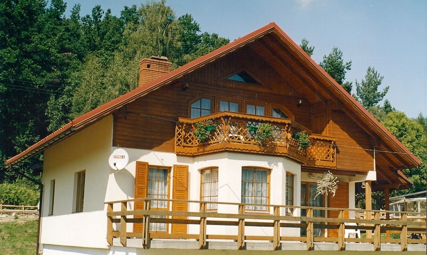 Dwukondygnacyjny dom z drewnianą elewacją na poddaszu, balkon z ozdobną balustradą i kwiatami, biała elewacja na parterze z brązowymi okiennicami, antena satelitarna na ścianie, otoczony zielenią.