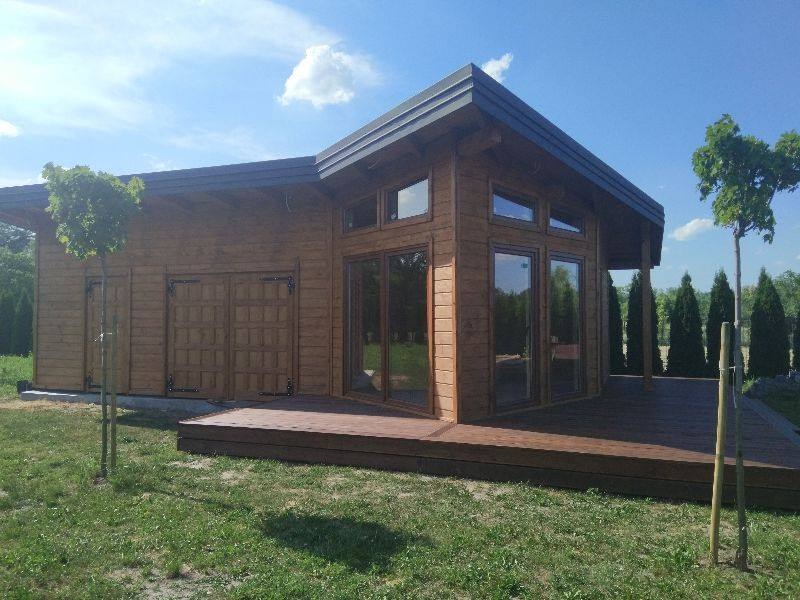 Gotowy, parterowy dom z drewna o nowoczesnej architekturze, z dużymi oknami i drewnianym tarasem, na tle zielonego trawnika i drzew.