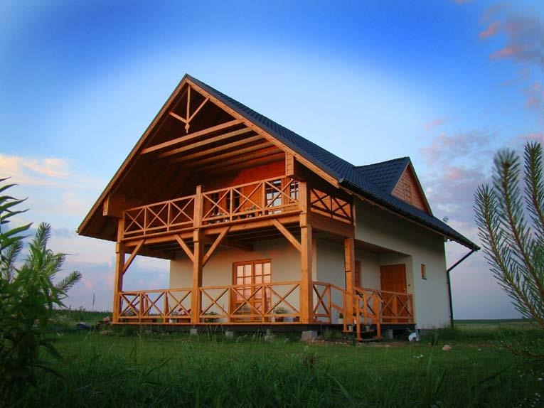 Gotowy dom szkieletowy z drewnianą konstrukcją dachu i balustradami, widoczny z poziomu trawy o zmierzchu, z fragmentem iglastego drzewa po prawej stronie.