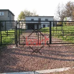 Nowoczesna, czarna brama wjazdowa z metalowymi prętami o geometrycznym wzorze, na tle żwirowego podjazdu i budynków.