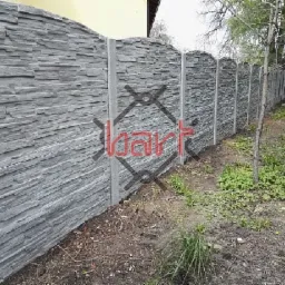 Szary, betonowy płot imitujący kamień łupany, z zaokrąglonymi g&oacute;rnymi krawędziami, ustawiony na betonowych słupkach, widoczny z bliska na tle drzew i trawy.