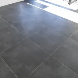 Nowo położone, duże, grafitowe płytki podłogowe w słonecznym pomieszczeniu z otwartym oknem; karton po zaprawie do fugowania stoi przy ścianie.