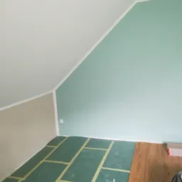 Układanie paneli podłogowych w pokoju ze skośnym sufitem, widoczna zielona mata wygłuszająca z taśmą klejącą i częściowo ułożone panele o wzorze drewna.