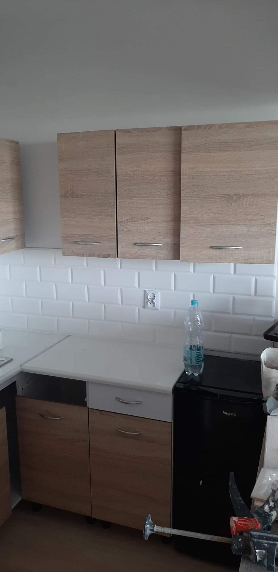 Kuchnia po remoncie: białe płytki typu metro na ścianie, jasne drewniane szafki wiszące i dolne, biały blat. Na blacie stoi butelka wody obok czarnej lodówki. Widoczne narzędzia malarskie na podłodze.