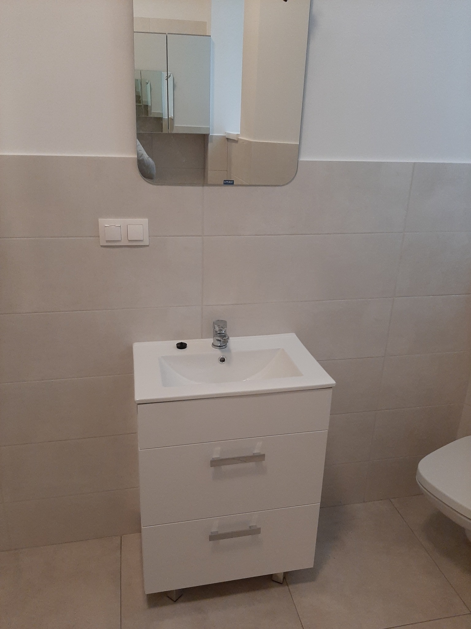 Biała umywalka z szafką i dwoma szufladami, chromowana bateria, beżowe płytki na ścianach i podłodze, lustro z szafką w tle, nowoczesna łazienka.