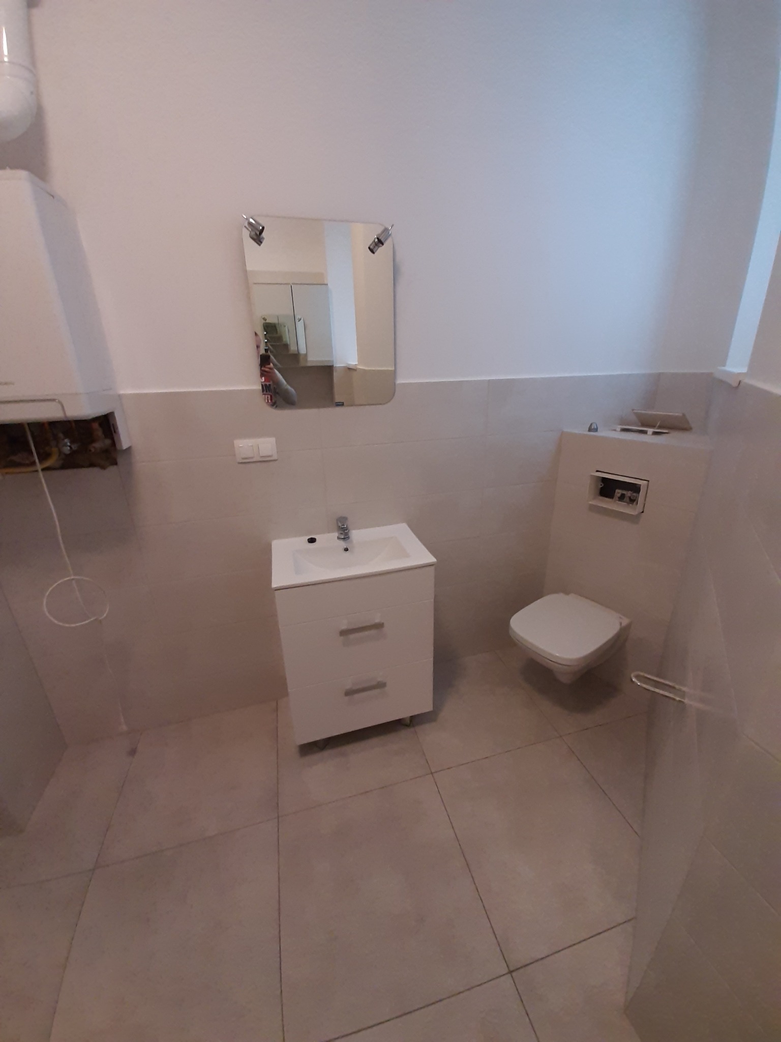 Minimalistyczna łazienka w trakcie remontu: biała umywalka z szafką, toaleta podtynkowa z otwartym panelem inspekcyjnym, widoczne płytki podłogowe i fragment bojlera z instalacją.