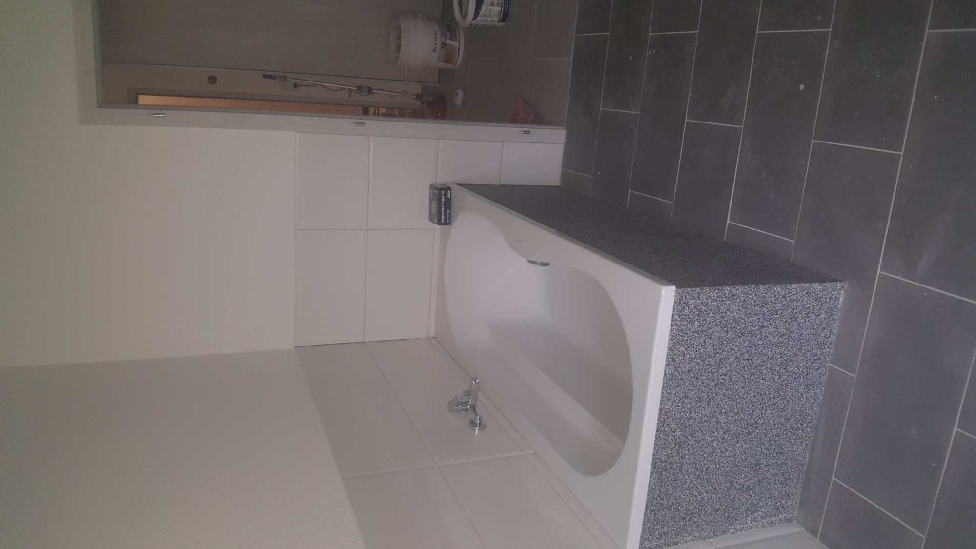 Wykończona łazienka z białą wanną obudowaną płytkami imitującymi lastryko, białe płytki ścienne, ciemnoszare płytki podłogowe, widoczna bateria wannowa.