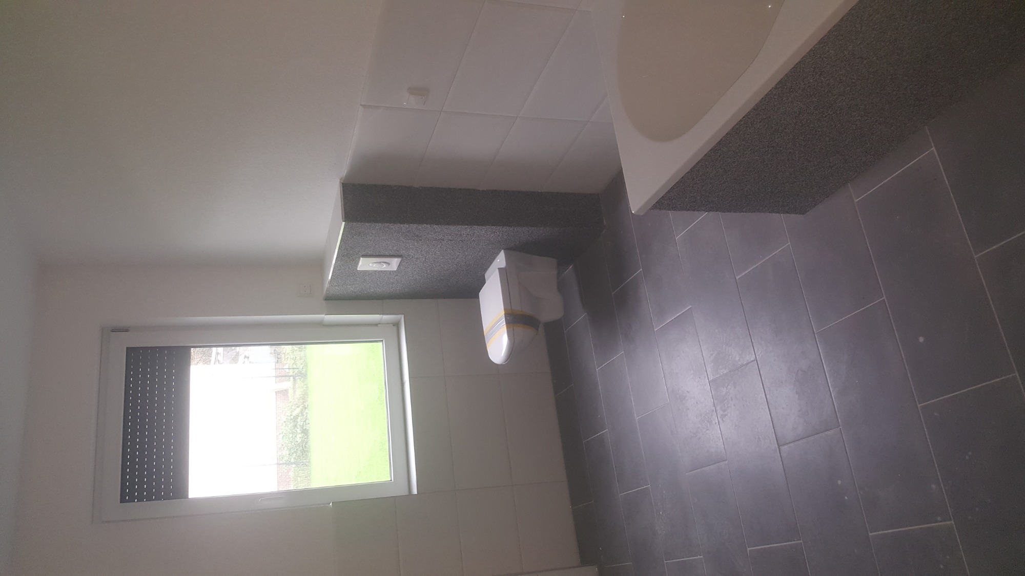 Wykończona łazienka z szarymi płytkami podłogowymi, białą glazurą ścienną, podwieszaną toaletą i oknem z roletą.