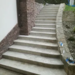 Betonowe schody zewnętrzne z kamienną obudową, prowadzące do ogrodu, widoczne narzędzia glazurnicze na krawędzi.