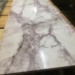 Płyta z białego marmuru z wyraźnymi szarymi i fioletowymi żyłami, leżąca na drewnianych paletach, oświetlona czerwoną linią lasera, wskazującą na precyzyjne cięcie lub pomiar.