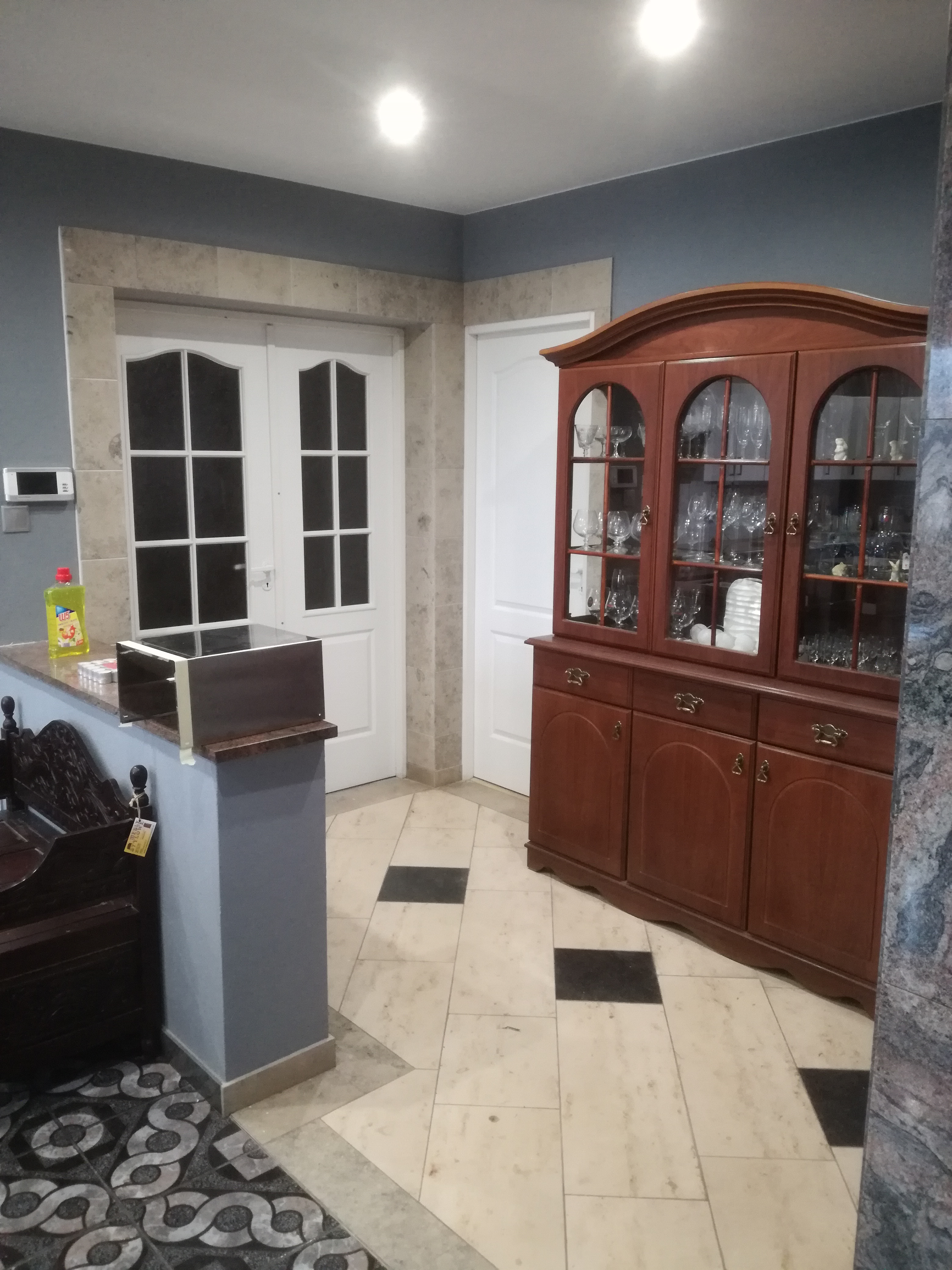 Wnętrze z kamienną ramą wokół białych drzwi, widoczna także drewniana witryna z szklanymi naczyniami i fragment podłogi z jasnych i ciemnych płytek.