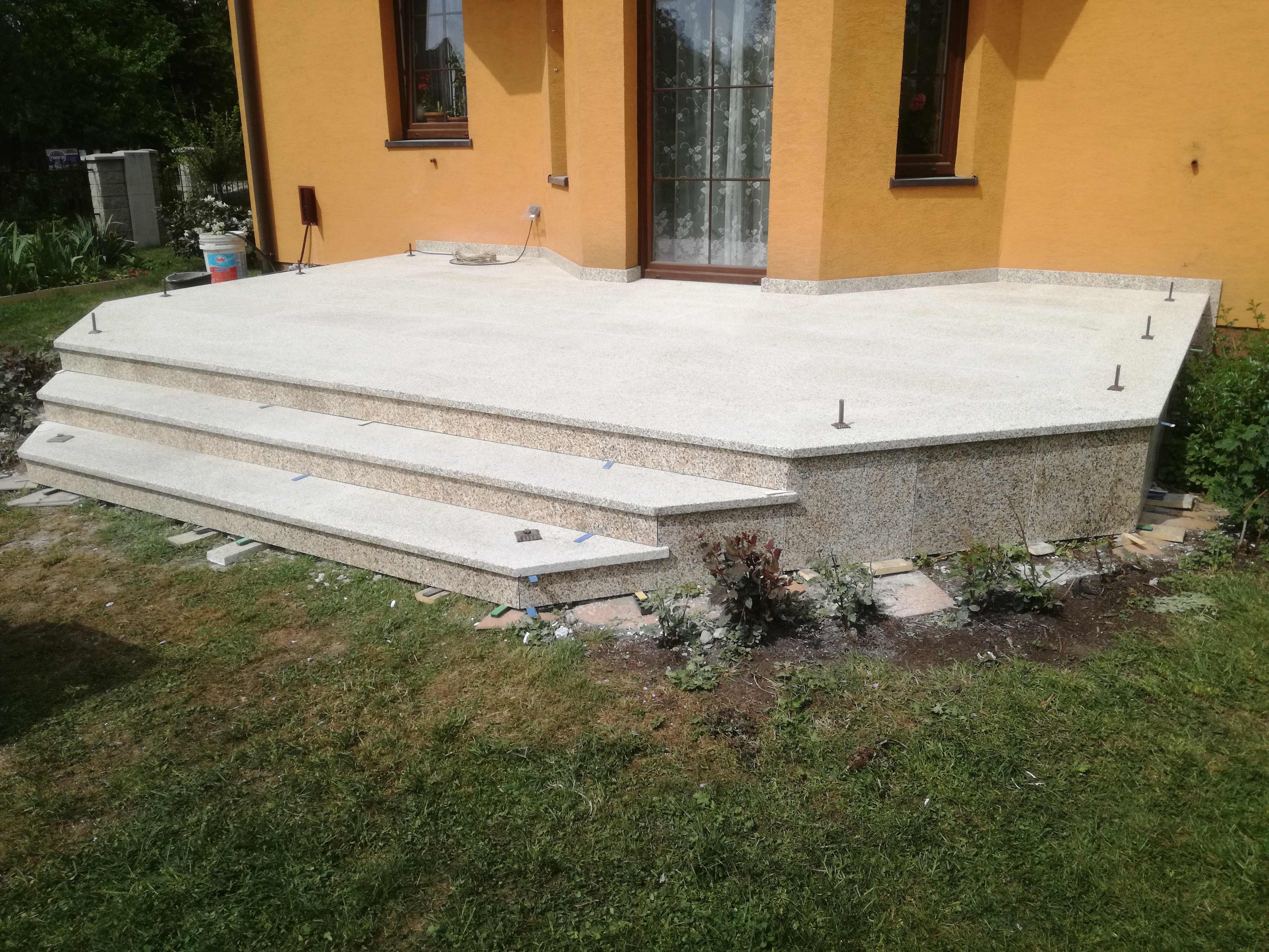 Jasny taras z trzema stopniami z materiału imitującego kamień, z widocznymi metalowymi elementami mocującymi, przylegający do pomarańczowego budynku z oknami i drzwiami.