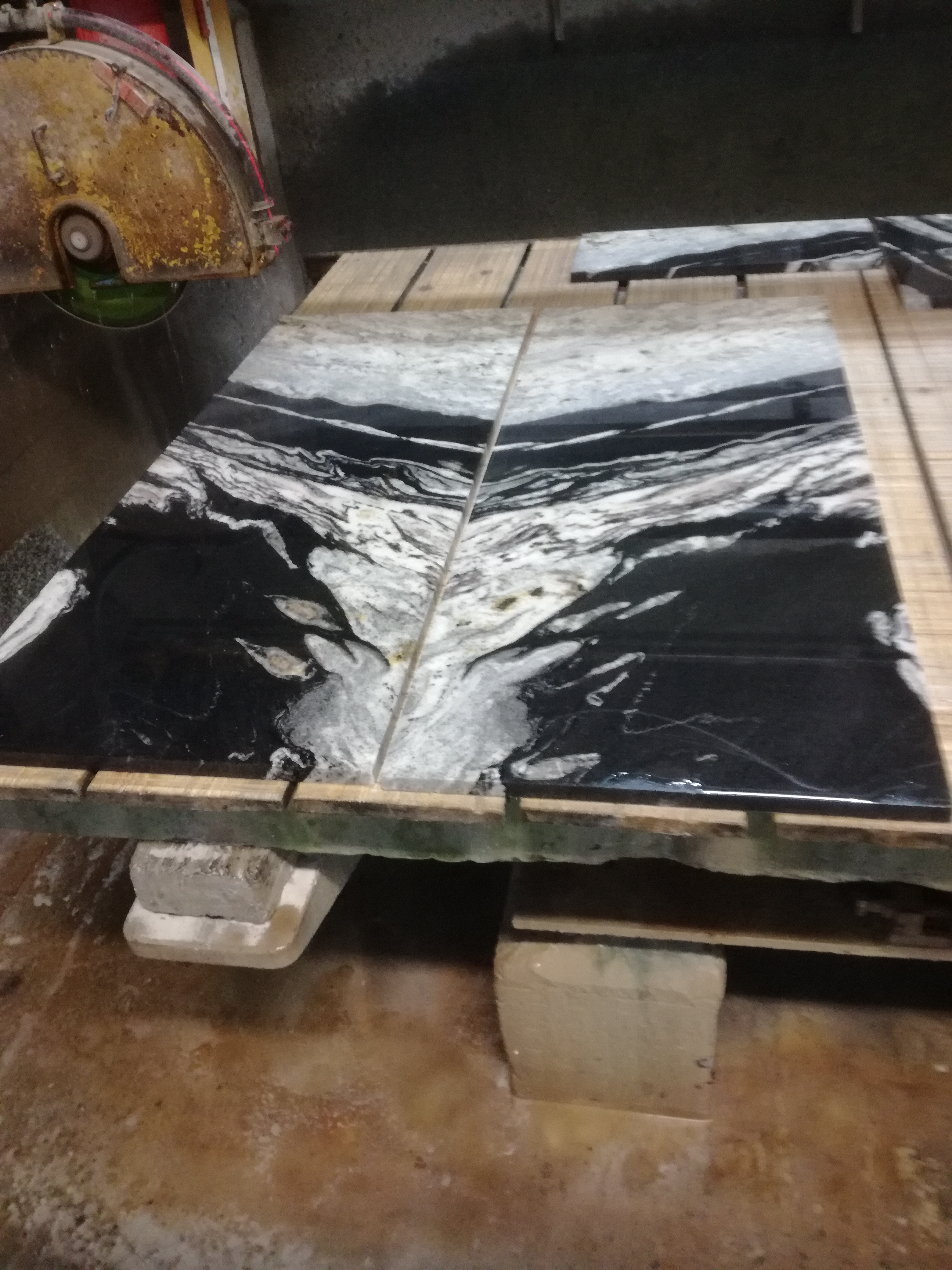 Dwa elementy blatu z czarno-białego kamienia z wyraźnym użyleniem, leżące na drewnianej palecie, obok maszyny do cięcia kamienia.