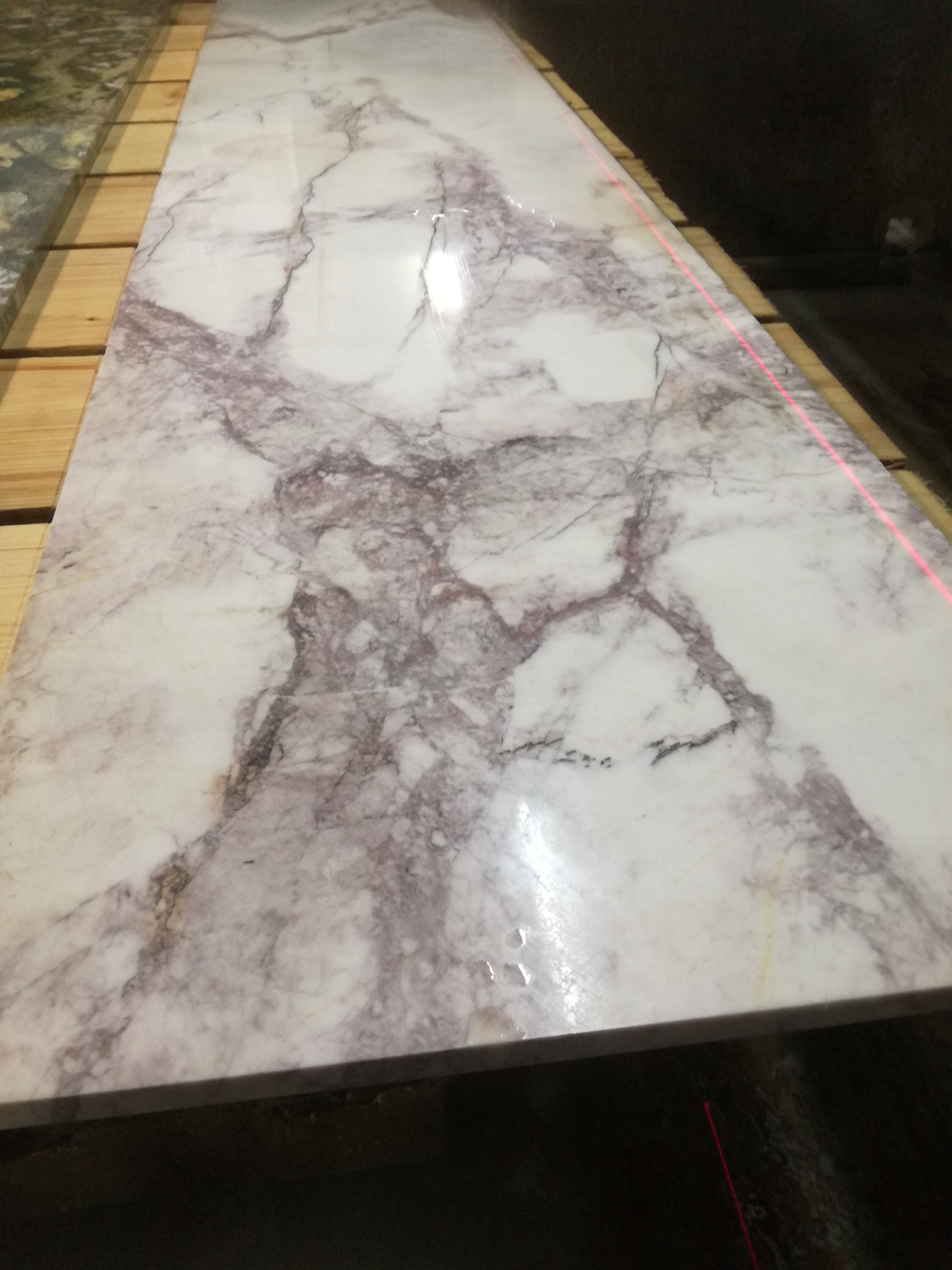 Płyta z białego marmuru z wyraźnymi szarymi i fioletowymi żyłami, leżąca na drewnianych paletach, oświetlona czerwoną linią lasera, wskazującą na precyzyjne cięcie lub pomiar.