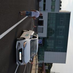 Nauka Jazdy SIS-Car - Biały samochód z literą 'L' na dachu, zaparkowany na placu manewrowym z pachołkami i instruktorem w tle, na tle nowoczesnego biurowca.