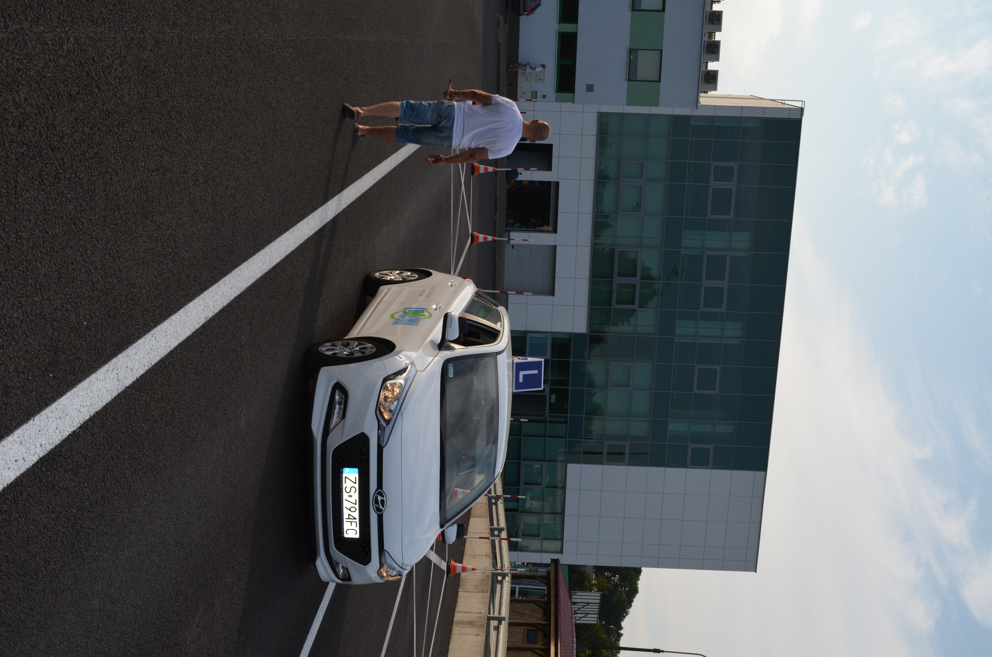 Biały samochód z literą 'L' na dachu, zaparkowany na placu manewrowym z pachołkami i instruktorem w tle, na tle nowoczesnego biurowca.