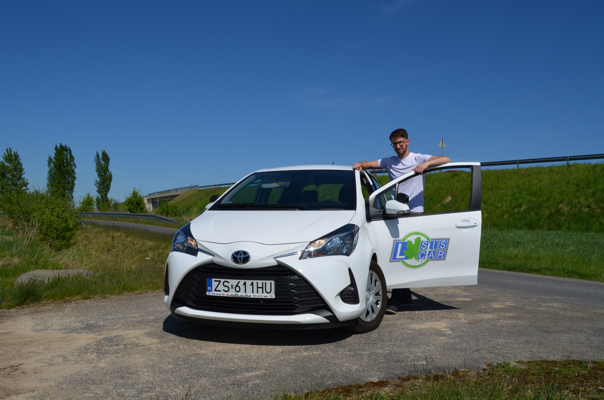 Biały samochód z otwartymi drzwiami, na których widnieje logo z napisem 'L SIS CAR', instruktor opiera się o dach pojazdu, w tle zielona trawa i błękitne niebo.