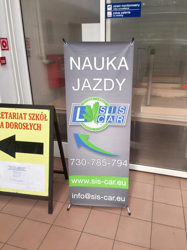 Rozkładany baner reklamowy szkoły jazdy SIS CAR z numerem telefonu i adresem strony internetowej, stojący obok tablicy informacyjnej sekretariatu szkół dla dorosłych.