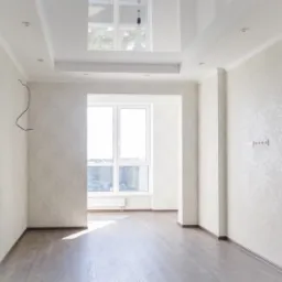 Jasne wnętrze po remoncie z panelami podłogowymi, tapetą w delikatny wzór i sufitem podwieszanym z oświetleniem punktowym; widoczne gniazdka elektryczne i wystające kable.