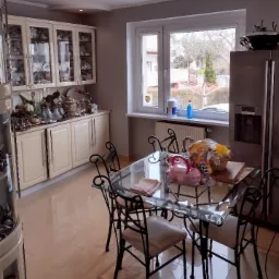 Jasna kuchnia z okrągłym sufitem podwieszanym z oświetleniem punktowym, kremowymi szafkami z przeszklonymi witrynami, szklanym stołem z kutymi krzesłami i błyszczącą podłogą z płytek.