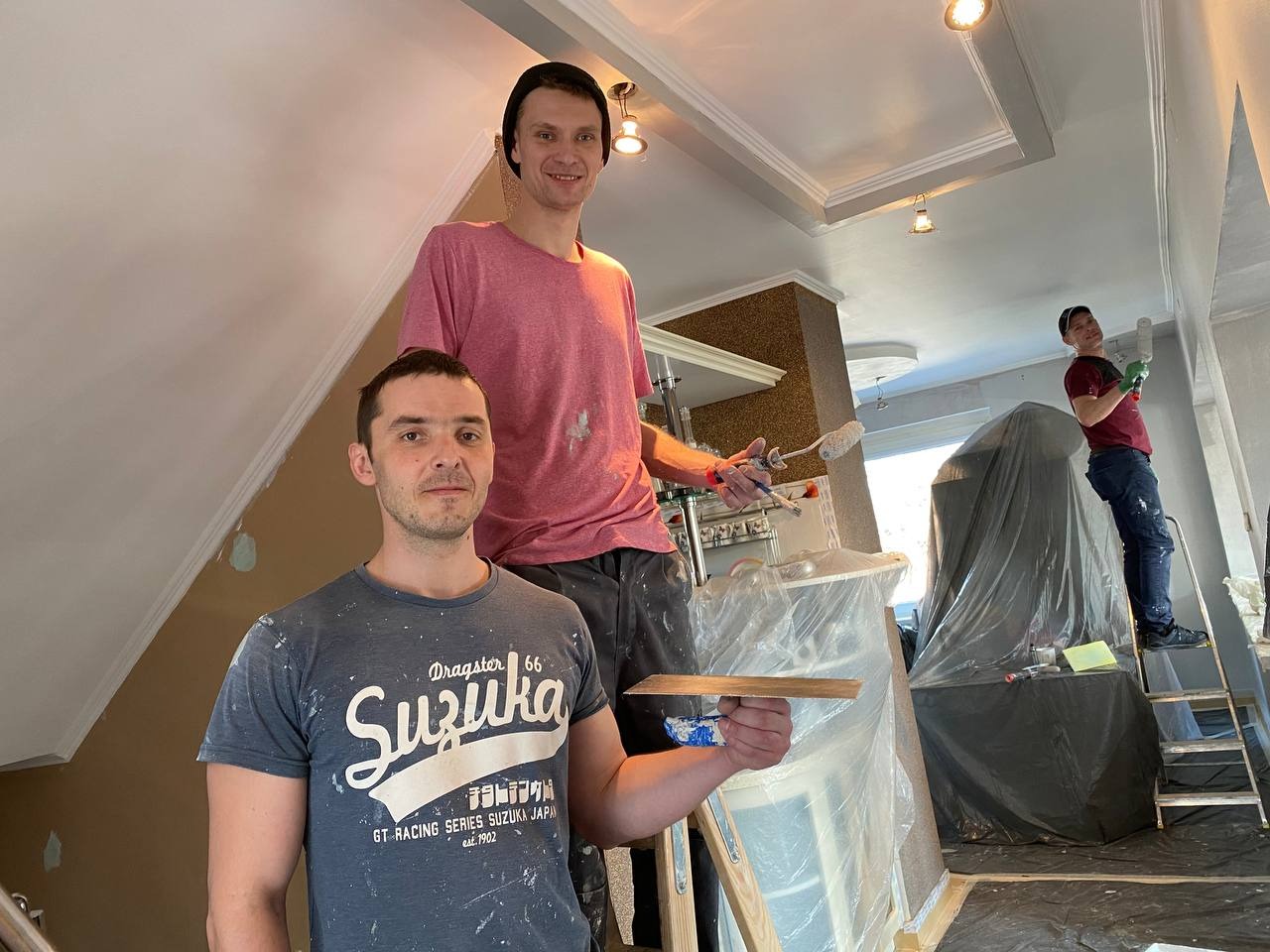 Trzech mężczyzn podczas malowania sufitu w pomieszczeniu z dekoracyjną sztukaterią, jeden na drabinie z wałkiem, drugi na podeście z pędzlem, trzeci z deską w dłoni, zabezpieczone meble folią.