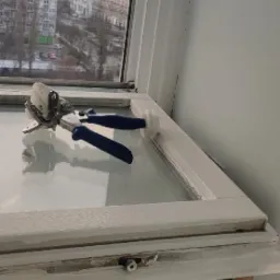 Szczypce do mocowania siatki moskitierowej leżące na ramie okiennej z widokiem na budynki w tle.