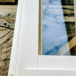 Fragment białej ramy okiennej z widoczną uszczelką i szybą, na tle ochronnej folii malarskiej.