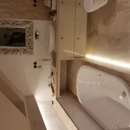 Nowoczesna łazienka z beżowymi płytkami imitującymi marmur, owalna wanna obudowana płytkami, biała umywalka z szafką i lustro w ozdobnej ramie.