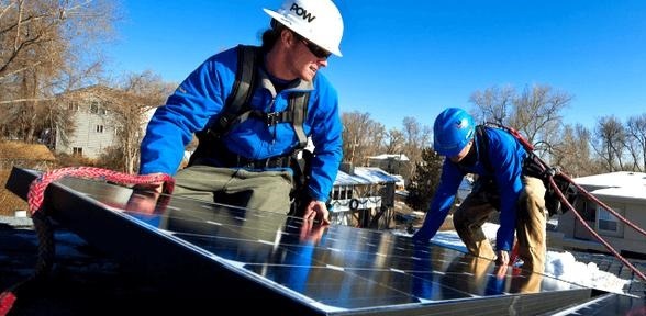 Dwóch monterów w kaskach i uprzężach zabezpieczających instaluje panele słoneczne na dachu pokrytym śniegiem w słoneczny dzień, z widocznymi domami w tle.