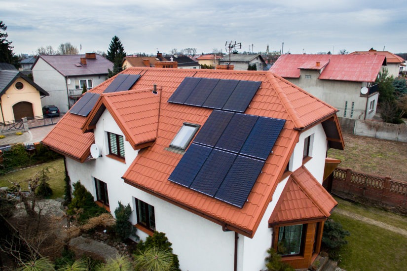 Widok z góry na dom z czerwoną dachówką, na której zamontowane są panele fotowoltaiczne, widoczna instalacja na tle innych domów i pochmurnego nieba.