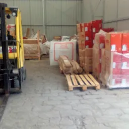 Wnętrze magazynu z żółtym wózkiem widłowym, paletami, paczkami styropianu oraz stosami kartonów zabezpieczonych folią.