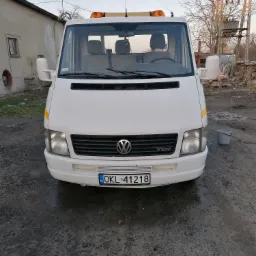 Biała laweta Volkswagen LT z pomarańczowymi światłami ostrzegawczymi na dachu, zaparkowana na betonowym placu przed budynkiem gospodarczym i słupem energetycznym.