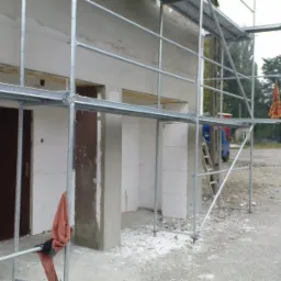 Budowa domu w stanie surowym zamkniętym z widocznymi rusztowaniami, ścianami z bloczków i wstępnie osadzonymi drzwiami; na rusztowaniu zwisa kolorowa szmatka.