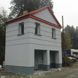 Dwukondygnacyjny, szary budynek w stanie surowym zamkniętym z pomarańczowym obramowaniem dachu i szarym cokołem, widoczne dwa otwory garażowe, obok leżą resztki styropianu.