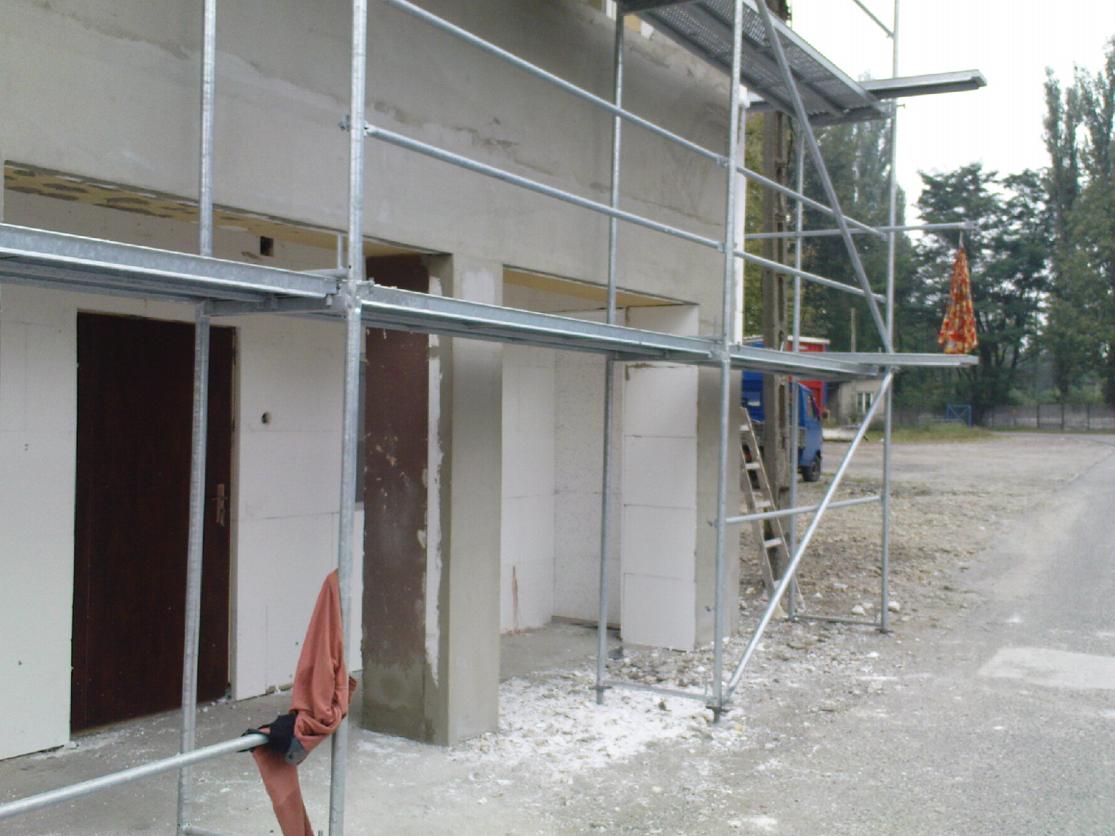 Budowa domu w stanie surowym zamkniętym z widocznymi rusztowaniami, ścianami z bloczków i wstępnie osadzonymi drzwiami; na rusztowaniu zwisa kolorowa szmatka.