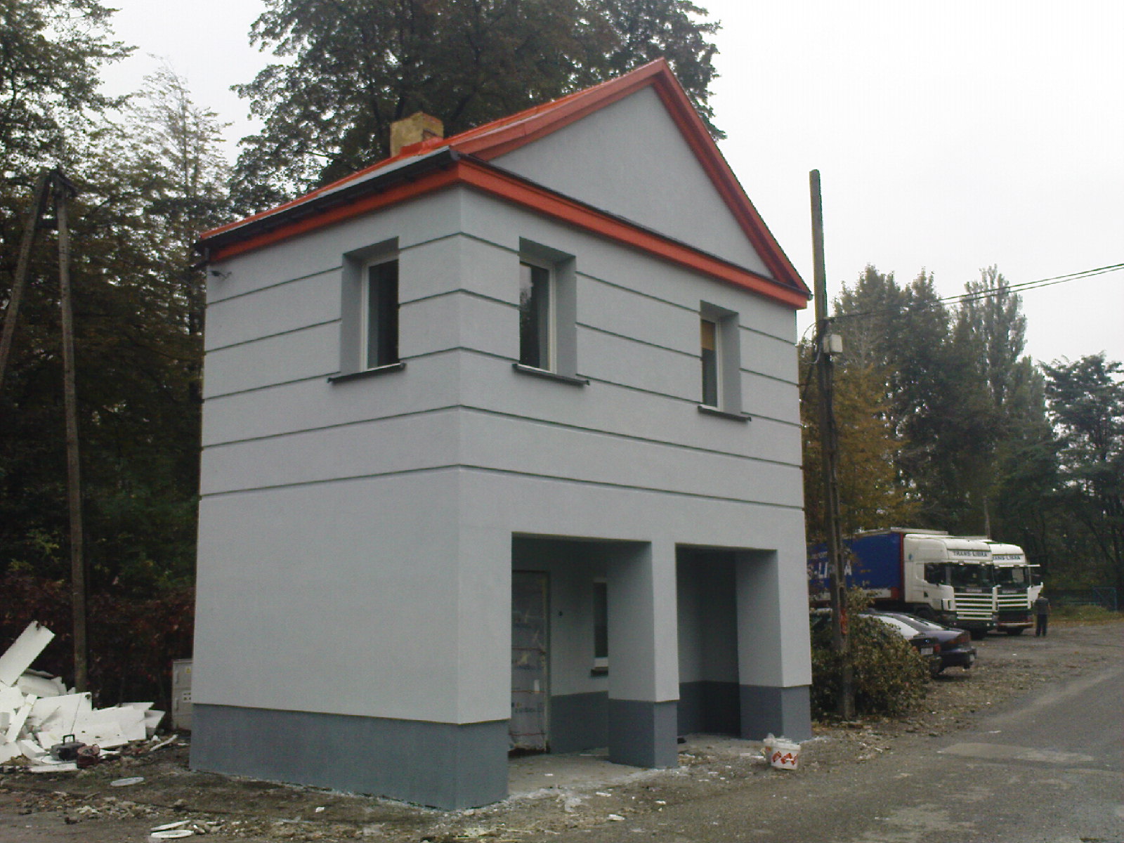 Dwukondygnacyjny, szary budynek w stanie surowym zamkniętym z pomarańczowym obramowaniem dachu i szarym cokołem, widoczne dwa otwory garażowe, obok leżą resztki styropianu.