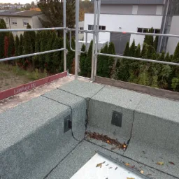 Szczegółowe ujęcie wykończonego, płaskiego dachu pokrytego szarą papą z widocznymi elementami mocującymi, wykonane z rusztowania w Grudziądzu, z tłem zielonych drzew i budynków.