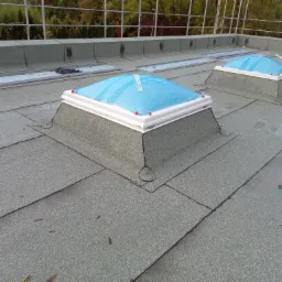 Widok na płaski dach pokryty papą z zamontowanymi świetlikami dachowymi zabezpieczonymi niebieską folią ochronną. W tle widoczne rusztowanie i drzewa.