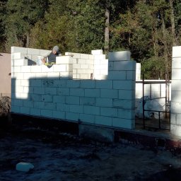 Talbud - Murarz w trakcie wznoszenia ścian parterowego domu z białych bloczków betonowych, widoczny fragment ściany sąsiedniego budynku i zalesione tło.