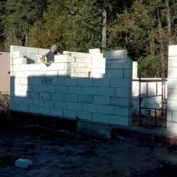 Murarz w trakcie wznoszenia ścian parterowego domu z białych bloczków betonowych, widoczny fragment ściany sąsiedniego budynku i zalesione tło.