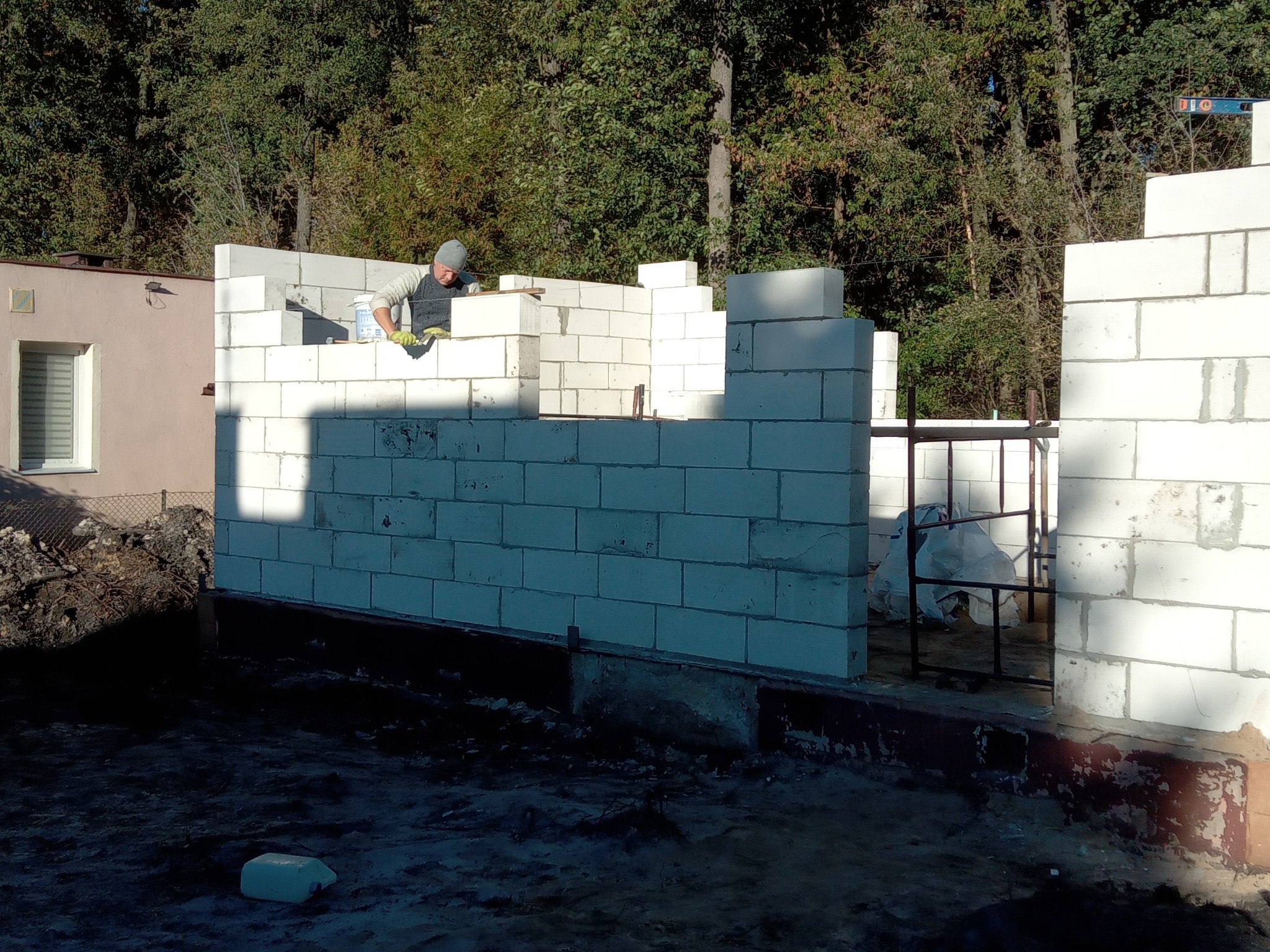 Murarz w trakcie wznoszenia ścian parterowego domu z białych bloczków betonowych, widoczny fragment ściany sąsiedniego budynku i zalesione tło.