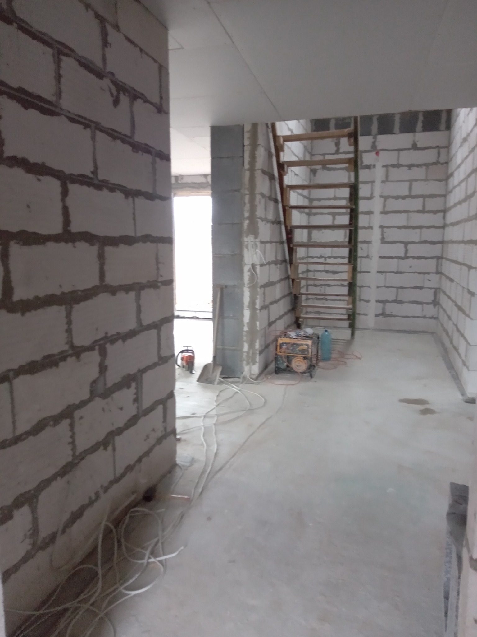 Wnętrze surowego domu w trakcie budowy z widocznymi ścianami z bloczków, drewnianą drabiną opartą o ścianę, generatorem prądu i plątaniną kabli na podłodze.