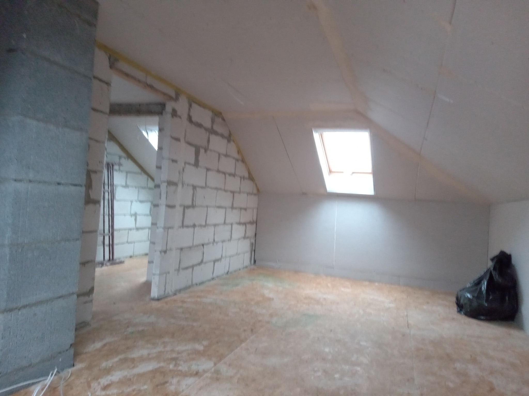 Poddasze w trakcie adaptacji: murowane ściany działowe z białego bloczka, okno dachowe, surowa podłoga z płyt OSB, czarny worek z odpadami w rogu.