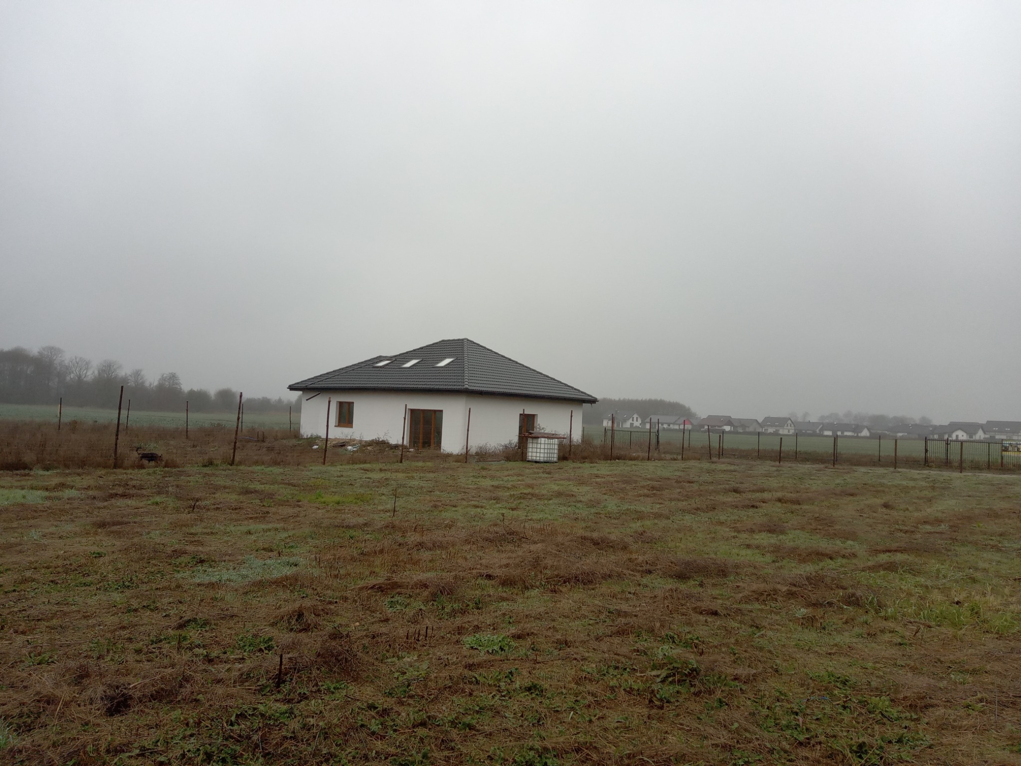 Nowo wybudowany, biały dom jednorodzinny z ciemną dachówką, otoczony metalowym ogrodzeniem na zarośniętej działce w mglisty dzień.