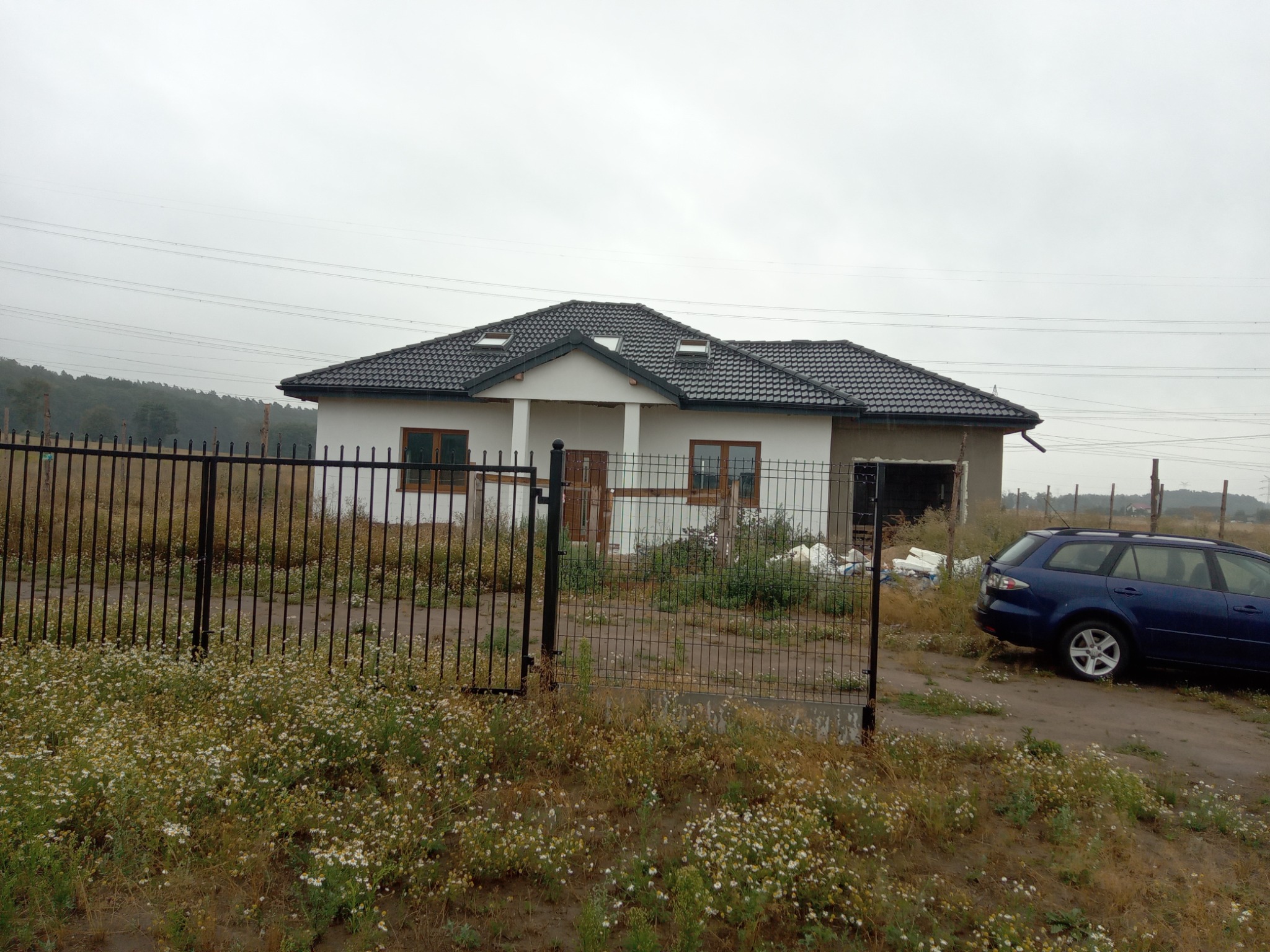 Niedokończony dom murowany z ciemną dachówką i oknami z brązowymi ramami, widoczny przez czarne metalowe ogrodzenie, z nieotynkowaną ścianą szczytową i zaparkowanym niebieskim kombi.