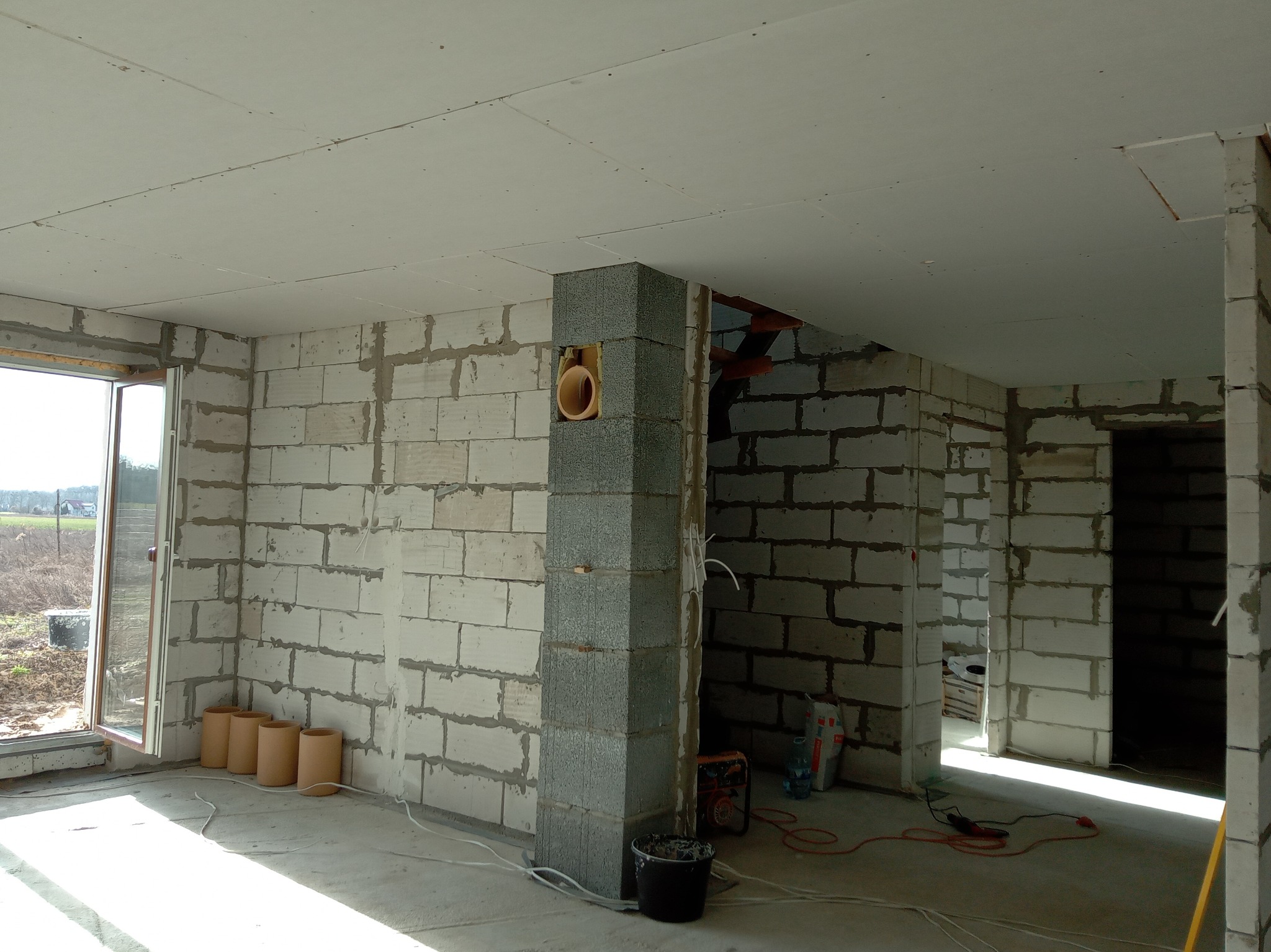 Wnętrze surowego domu w trakcie budowy: ściany z betonu komórkowego, sufit z płyt gipsowo-kartonowych, pion wentylacyjny z otworem na rurę, okno balkonowe z widokiem na zewnątrz, narzędzia...